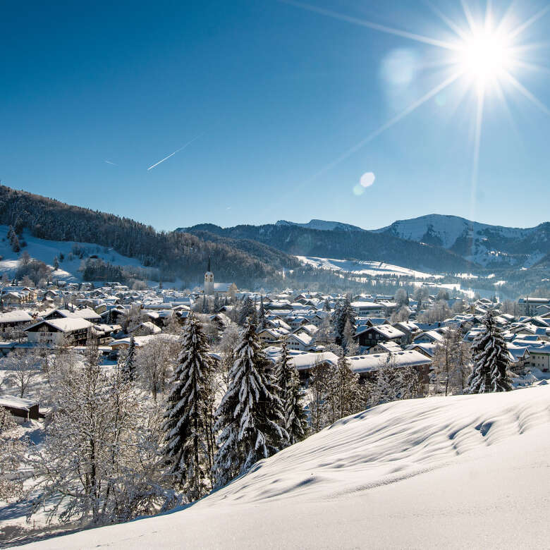 Blick auf den schneebedeckten Ortskern von Oberstaufen und das Bergpanorama der Nagelfluhkette im Schnee an einem sonnigen Wintertag.
