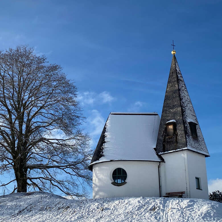 Verschneite Bruder Klaus Kapelle in Hagspiel Alte Kapelle auf einer verschneiten Wiese, daneben ein kahler Baum.