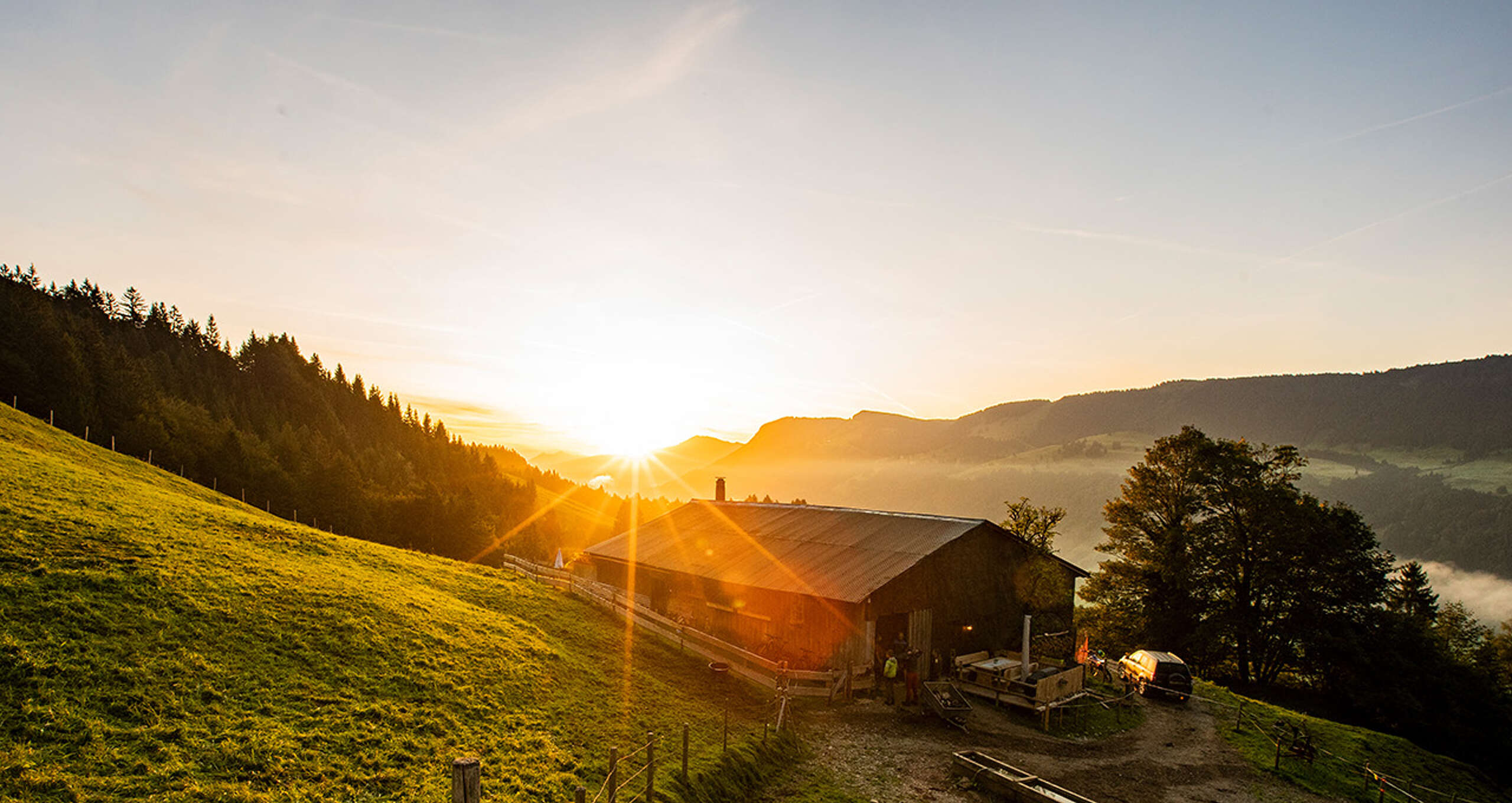 Blick Richtung Sonnenaufgang, davor eine Alpe und grüne Wiesen.