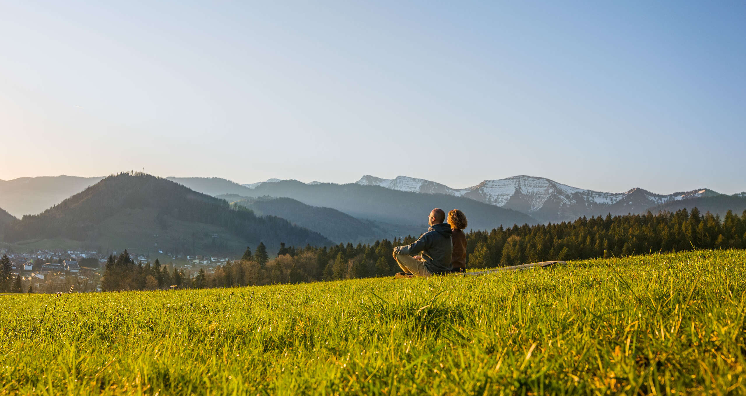 Mann und Frau sitzen in Wiese mit Blick aauf Oberstaufen und die umliegenden Berge bei Sonnenaufgang