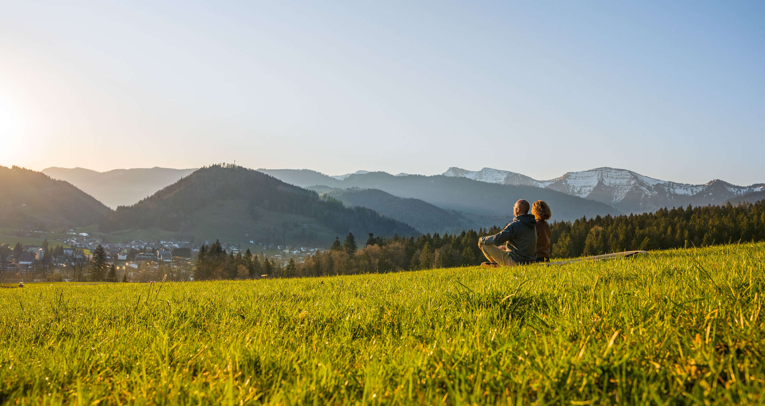 Mann und Frau sitzen in Wiese mit Blick aauf Oberstaufen und die umliegenden Berge bei Sonnenaufgang