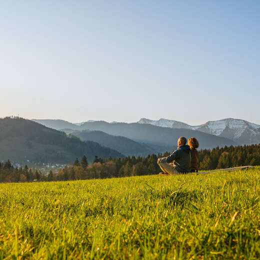 Mann und Frau sitzen in Wiese mit Blick aauf Oberstaufen und die umliegenden Berge bei Sonnenaufgang