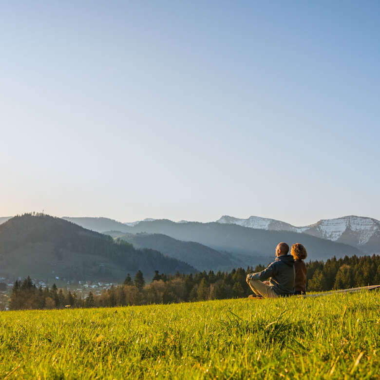 Mann und Frau sitzen in Wiese mit Blick aauf Oberstaufen und die umliegenden Berge bei Sonnenaufgang
