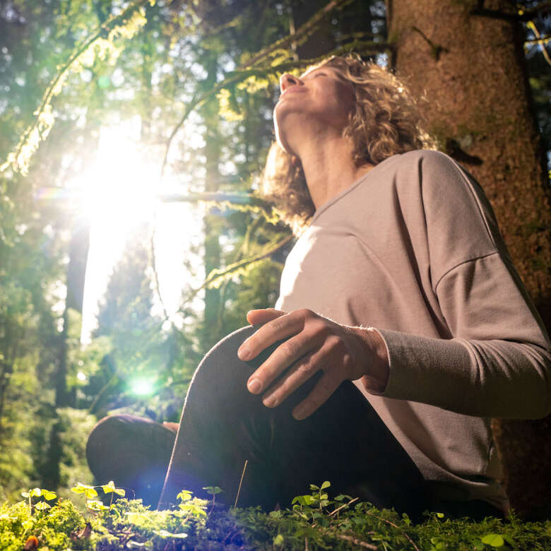 Den Wald mit allen Sinnen spüren Frau lehnt an einem Baum im Wald in der Sonne