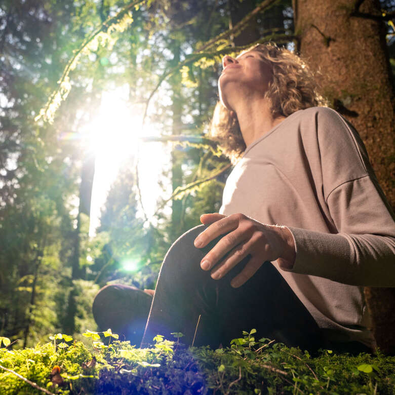 Den Wald mit allen Sinnen spüren Frau lehnt an einem Baum im Wald in der Sonne
