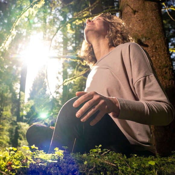 Den Wald mit allen Sinnen spüren Frau lehnt an einem Baum im Wald in der Sonne