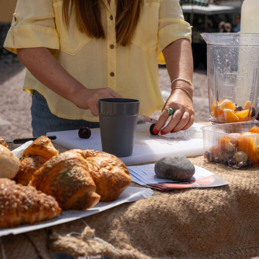 Person bereitet frische Smoothies mit Obst auf einem Outdoor-Tisch zu, daneben Croissants und Gebäck.