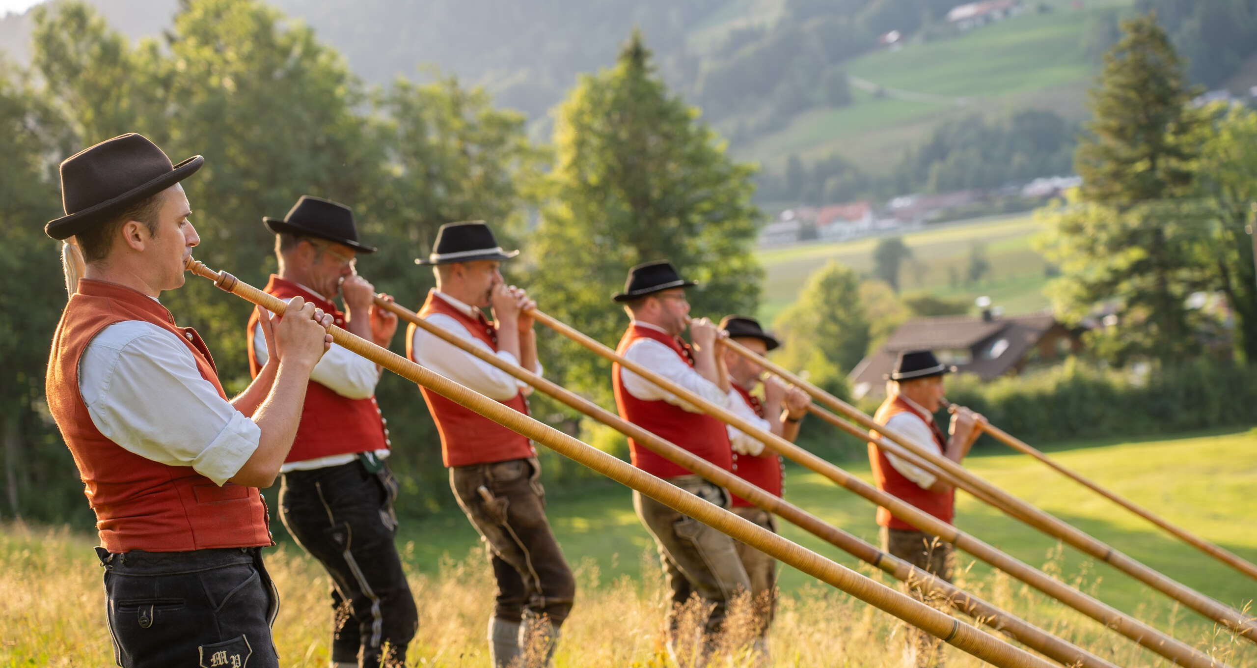 Alphornbläser in Tracht vor Bergkulisse in Oberstaufen Alt-Text: Alphornbläser in traditioneller Allgäuer Tracht mit Trachtenhut und roter Weste blasen Alphörner auf einer Alm bei Oberstaufen im Allgäu
