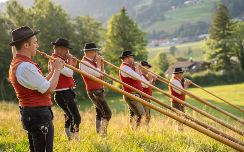 Alphornbläser in traditioneller Allgäuer Tracht mit Trachtenhut und roter Weste blasen Alphörner auf einer Alm bei Oberstaufen im Allgäu