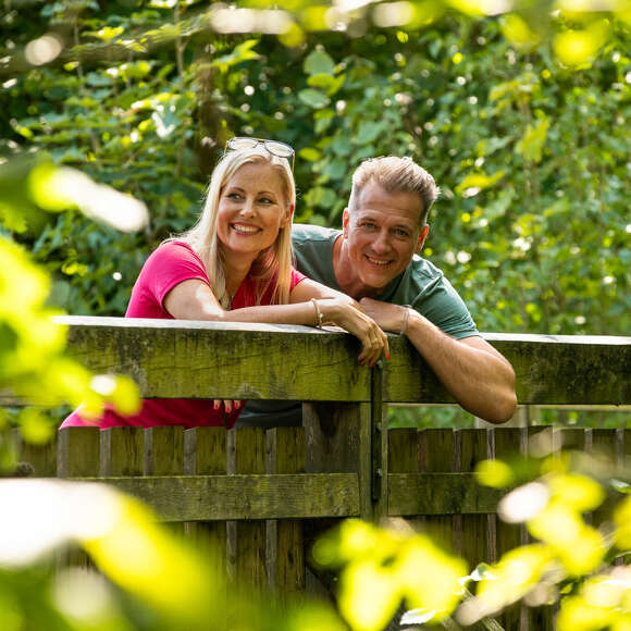 Mann und Frau lehnen an einem Holzsteg und lachen umrahmt von Blättern.