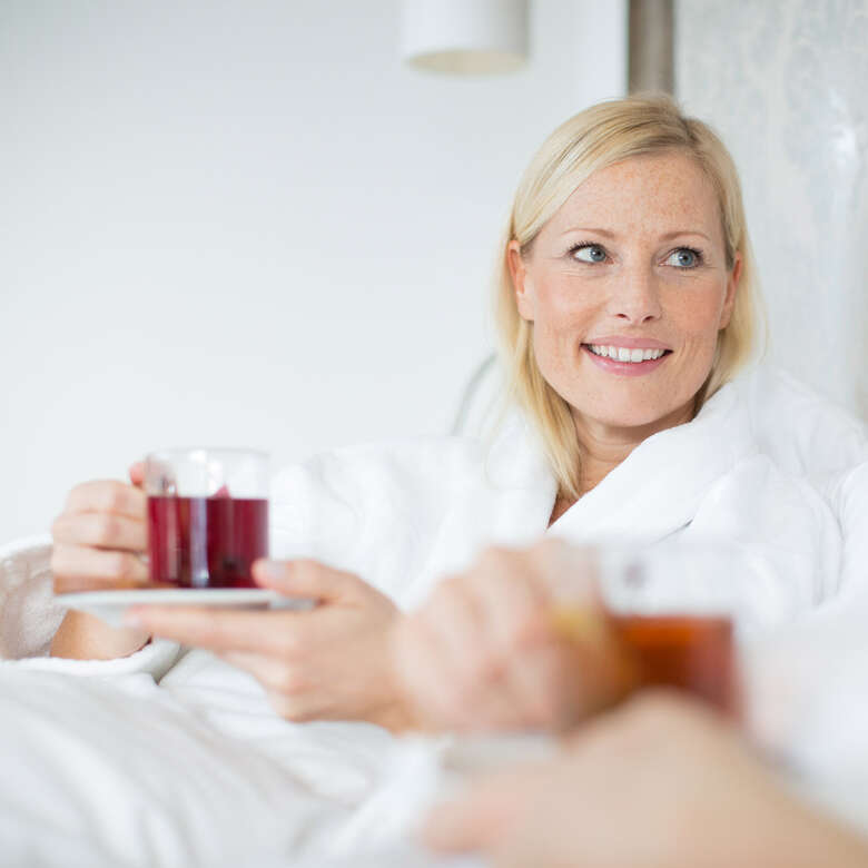Frau im Bademantel sitzt angelehnt im Bett mit einem Glas Tee in den Händen.