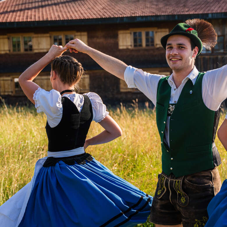 Tracht und Tradition – Oberstaufener Brauchtum bei den Huimatwochen Trachtengruppe beim traditionellen Volkstanz in Oberstaufen – Männer in grüner Weste und Frauen im Dirndl tanzen auf einer Wiese