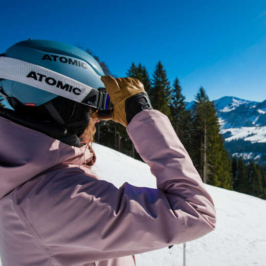 Skifahrerin fasst sich an die Skibrille und schaut in die Ferne.
