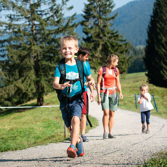 Fröhliche Familie wandert bei Sonnenschein am Hündle bei Oberstaufen im Allgäu – vorne ein lachender Junge mit Rucksack