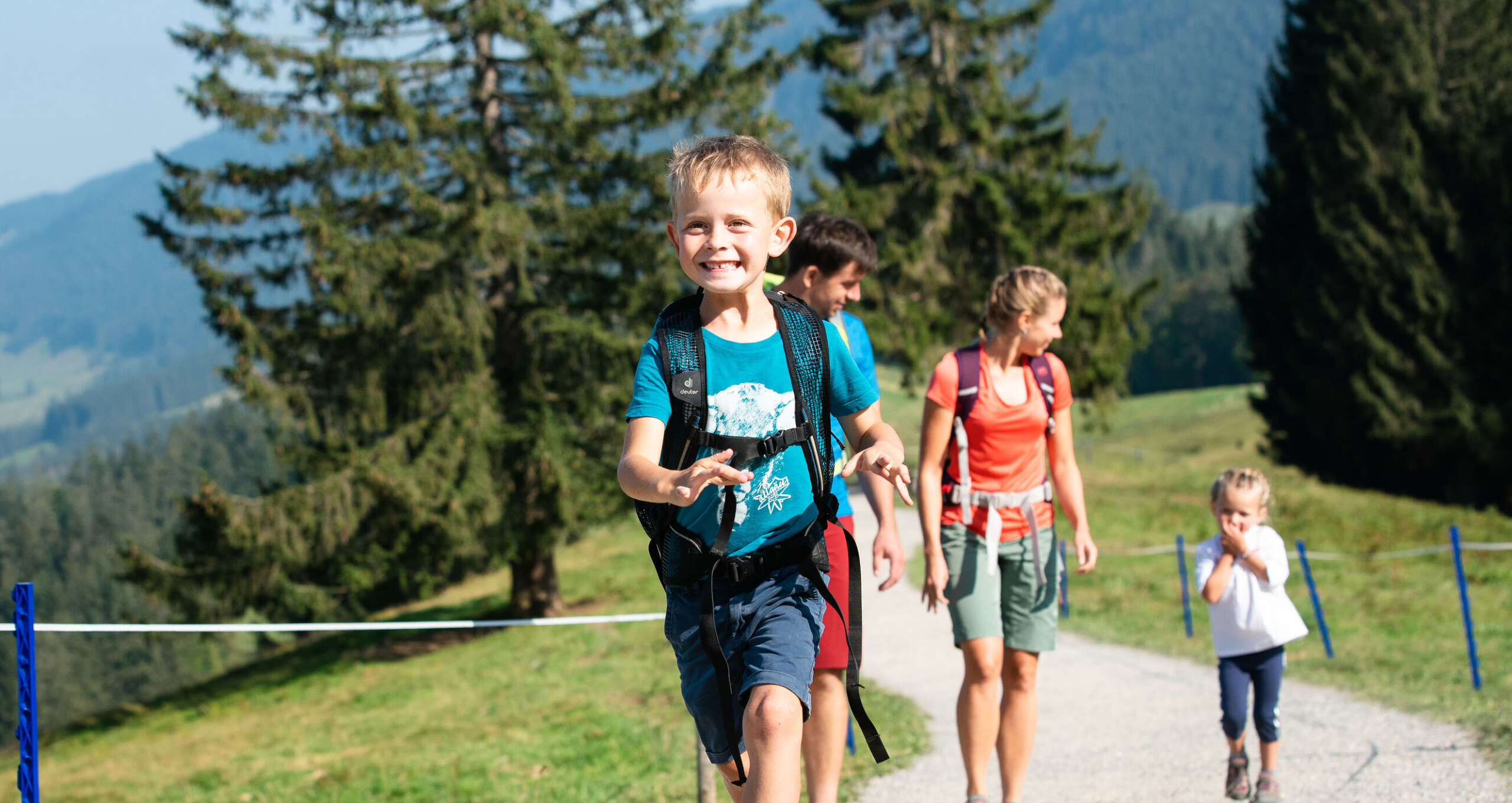 Fröhliche Familie wandert bei Sonnenschein am Hündle bei Oberstaufen im Allgäu – vorne ein lachender Junge mit Rucksack