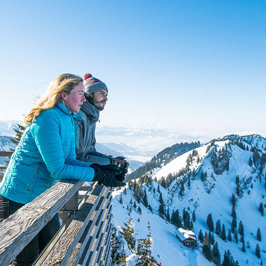 Paar lehnt an einer Brüstung in den Bergen mit verschneitem Bergpanorama an einem sonnigen Wintertag.