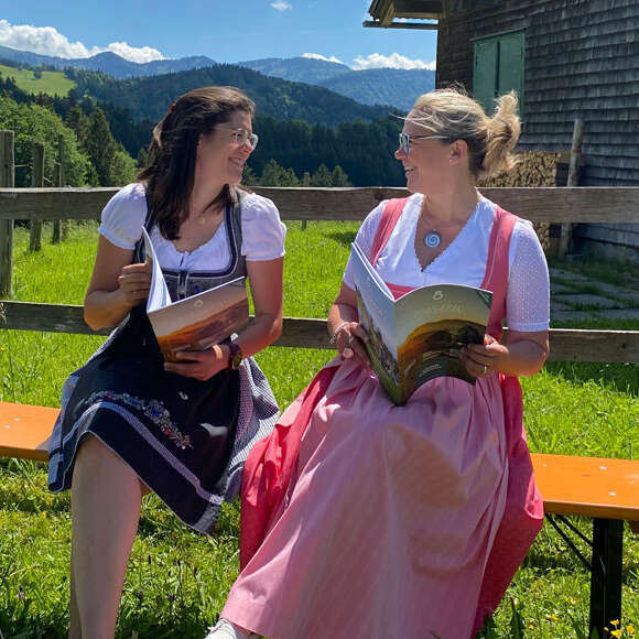 Zwei Frauen im Dirndl lesen in der Magazin Ausgabe Sommer 2022 vor einer Hütte mit Bergpanorama.