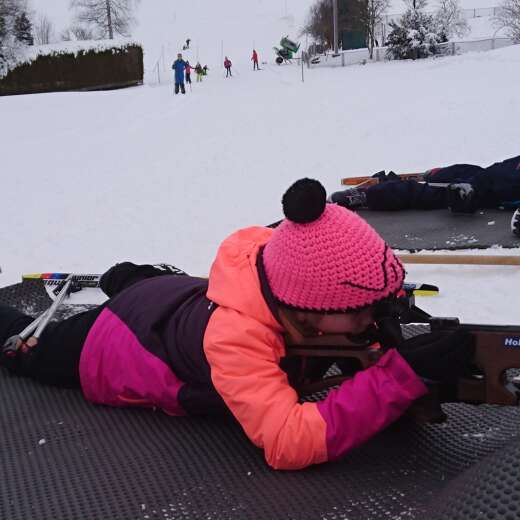 Kind mit Biathlongewehr im liegenden Anschlag auf der Loipe.