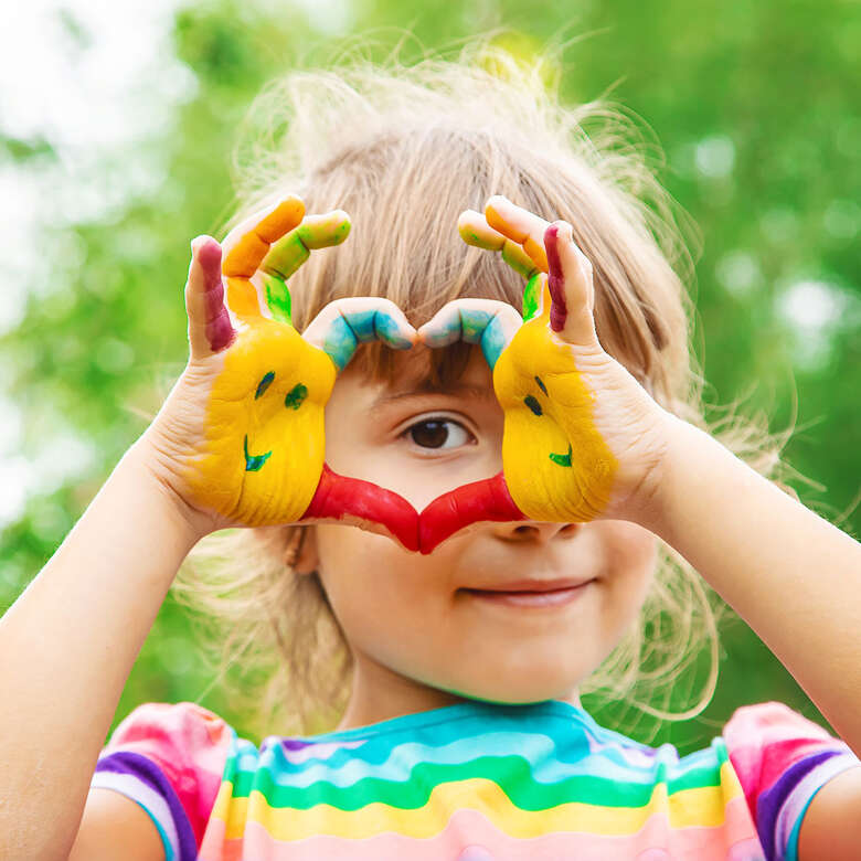 Kindersommer 2025 in Oberstaufen – Farbenfrohes Titelbild für Familienurlaub im Allgäu Titelbild mit fröhlichem Kind, das durch bunte, bemalte Hände schaut – Kindersommer 2025 in Oberstaufen im Allgäu