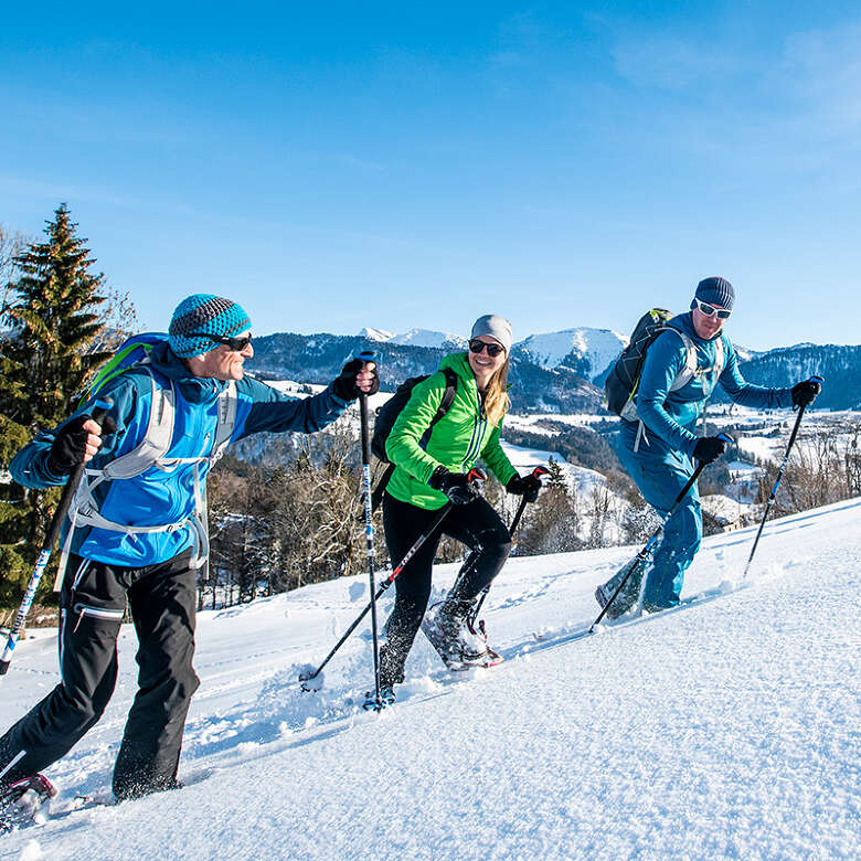 Drei Schneeschuhgeher über Oberstaufen mit dem verschneiten Hochgratpanorama.