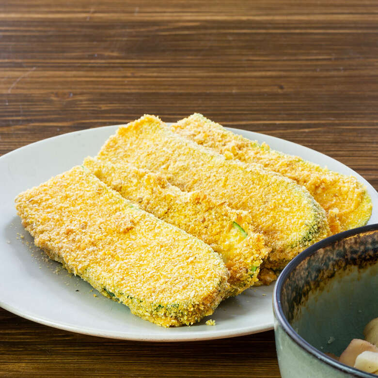 Auf einem weißen Teller liegen vier Scheiben Zucchini, paniert in glutenfreien Cornflakes.
