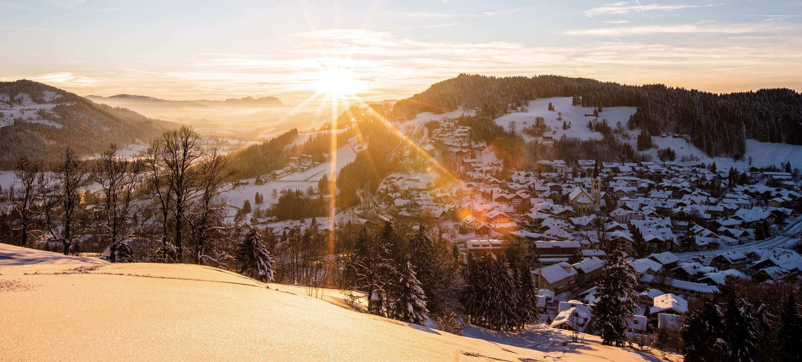 Sonne geht hinter dem verschneiten Ort von Oberstaufen unter.