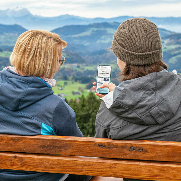 Frauen sitzen auf einer Bank mit Bergblick und der digitalen Tagesübersicht auf dem Handy