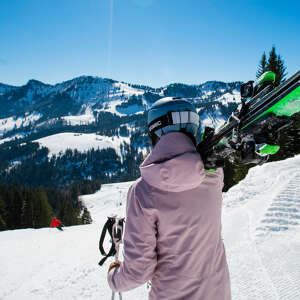 Frau im Skioutfit hält ihre Ski auf der Schulter und blickt in die verschneite Natur.