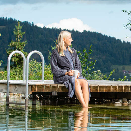 Frau sitzt im Bademantel auf einem Holzsteg an einem Schwimmteich. Sie blick in die Ferne, im Hintergrund grüne Natur.