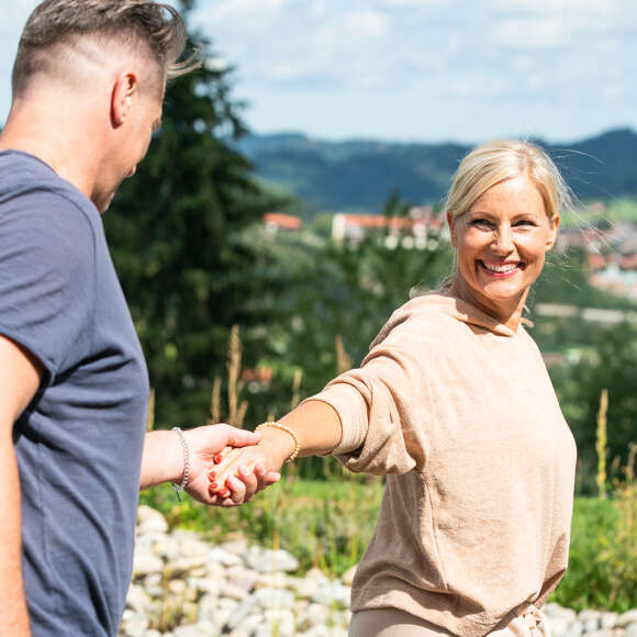 Mann und Frau sind draußen in einem Garten und die Frau zieht den Mann an der Hand zu sich.