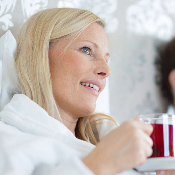 Frau sitzt im Bett und hält ein Glas mit roten Tee in der Hand.