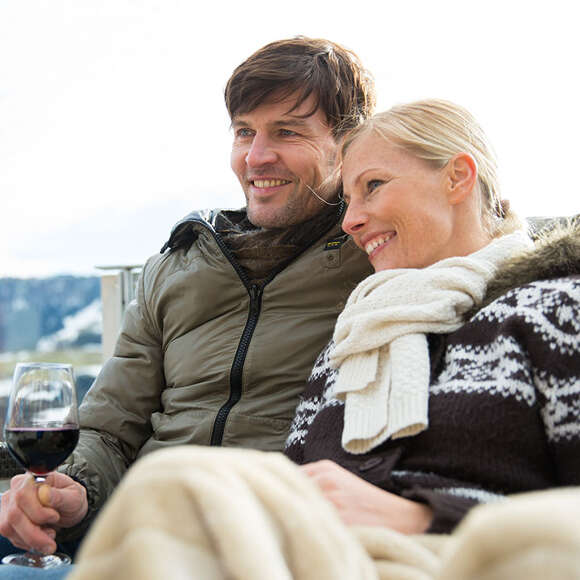 Mann und Frau in Winterjacken sitzen zusammengekuschelt in der Natur.