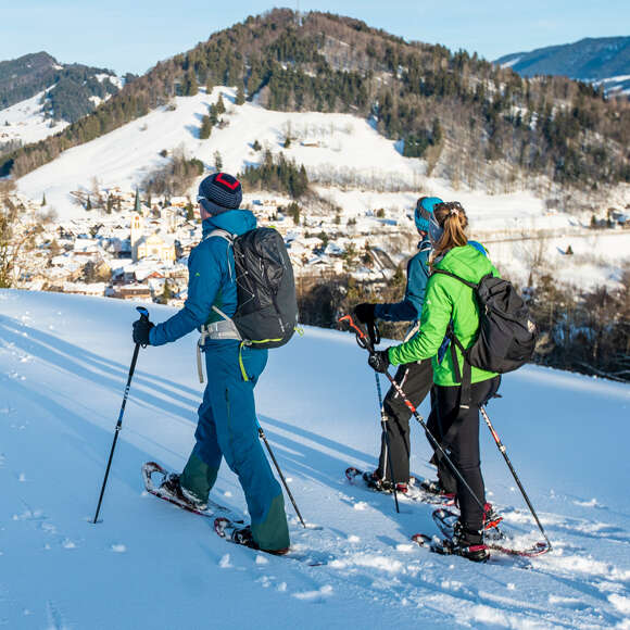 Drei Schneeschuhwanderer laufen durch den Schnee mit Blick auf Oberstaufen Ort.