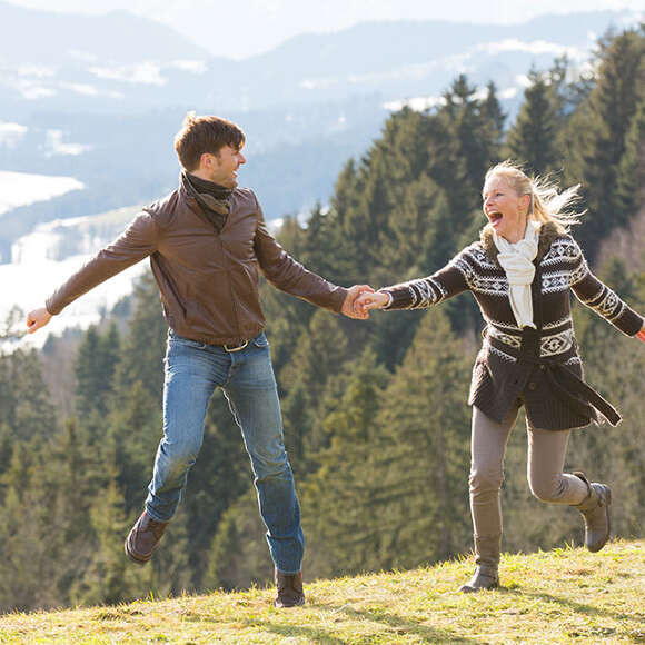 Mann und Frau tanzen lachend auf einem Hügel im Herbst. Im Hintergrund der erste Schnee.