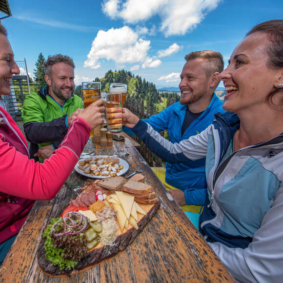 Vier Freunde sitzen auf einer Biergarnitur auf einer Terrasse in den Bergen und stoßen mit Getränken an. Auf dem Tisch steht eine Platte mit Wurst, Käse und Brot.
