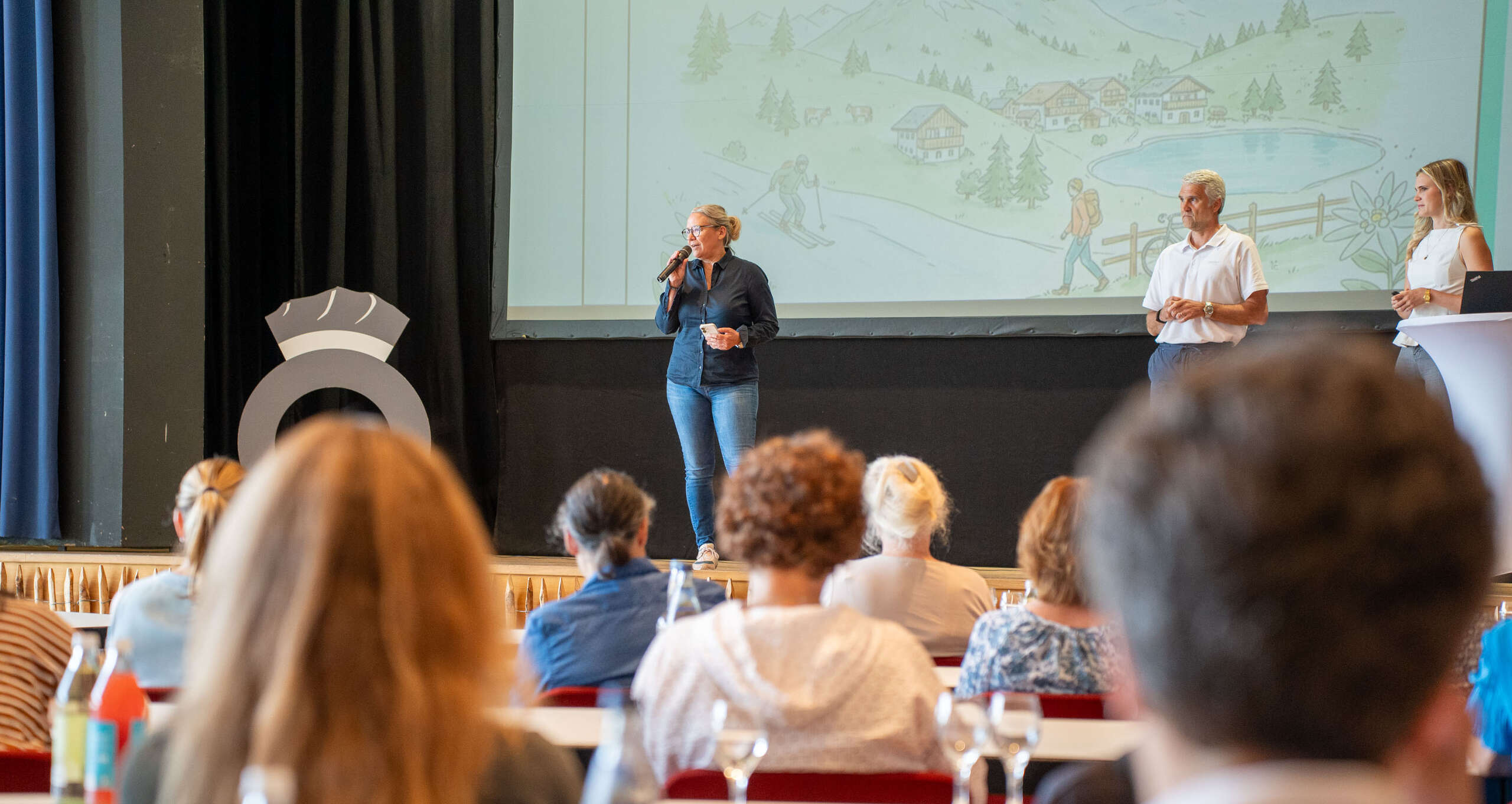 Netzwerkveranstaltung für touristische Leistungsträger Zwei Referentinnen und ein Mann auf einer Bühne sprechen vor sitzendem Publikum bei einer Tourismus-Schulung.