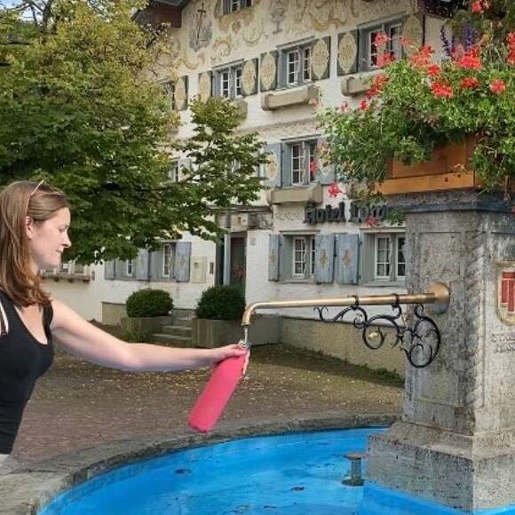Frau hält pinke Trinkflasche unter Wasserauslauf in Brunnen.