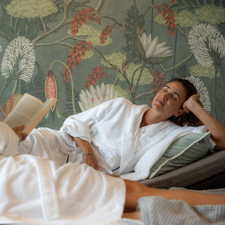 Gemeinsames Lesen in entspannter Atmosphäre Zwei Personen liegen entspannt auf einem Sofa vor einer floralen Tapete; eine Person liest in einem Buch, während die andere sich zurücklehnt.