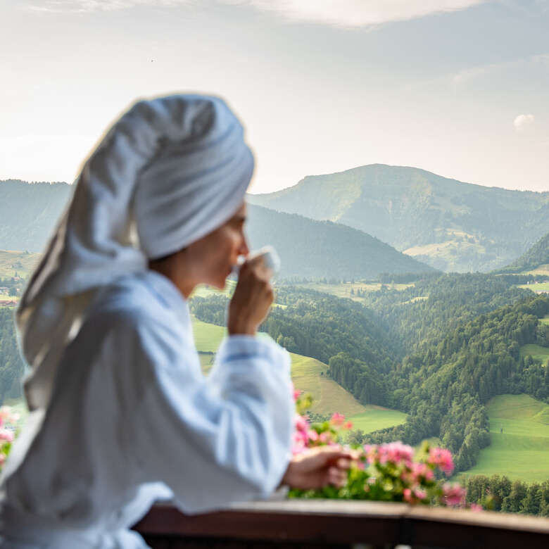 Wellness und Erholungsurlaub Frau im Bademantel mit Handtuchturban blickt von einer Terrasse in Oberstaufen auf die Allgäuer Hügellandschaft.
