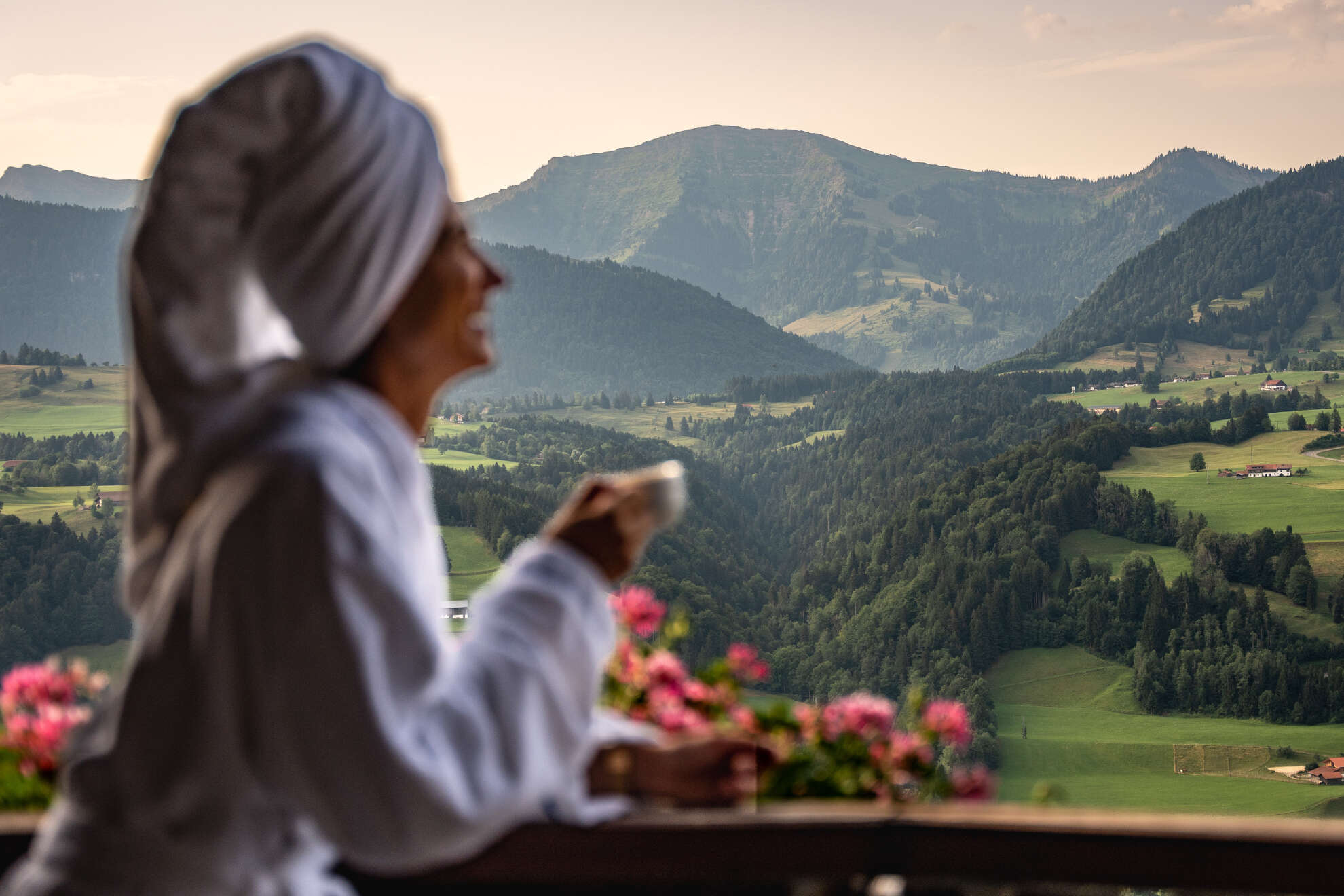 Wellness und Erholungsurlaub Frau im Bademantel mit Handtuchturban blickt von einer Terrasse in Oberstaufen auf die Allgäuer Hügellandschaft.