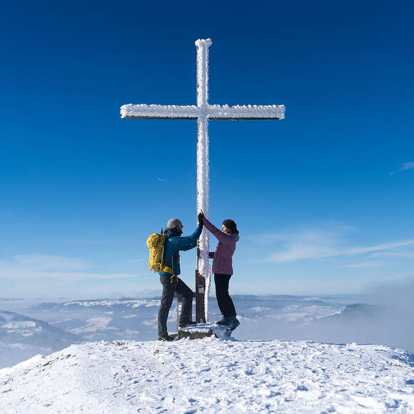 Wanderer am verschneiten Gipfelkreuz des Hündle mit Ausblick auf die weiße Landschaft von Oberstaufen