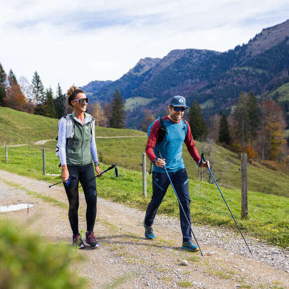 Wandern im Allgäu – Panoramawege Zwei Wanderer mit Stöcken auf einem sonnigen Panoramaweg im Allgäu, im Hintergrund grüne Berge.