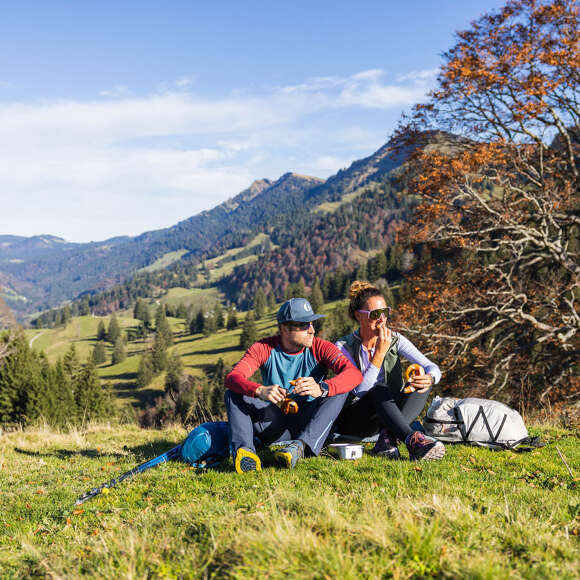Ein Pärchen sitzt auf der Kuppe und genießt die Sonne. Im Hintergrund sind Berge und herbstliche Wälder zu sehen.