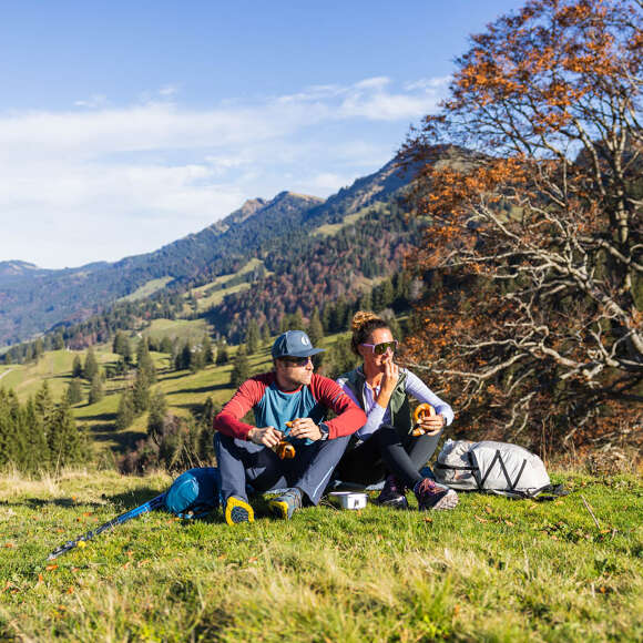 Wandern im Herbst bei Oberstaufen im Allgäu Ein Pärchen sitzt auf der Kuppe und genießt die Sonne. Im Hintergrund sind Berge und herbstliche Wälder zu sehen.