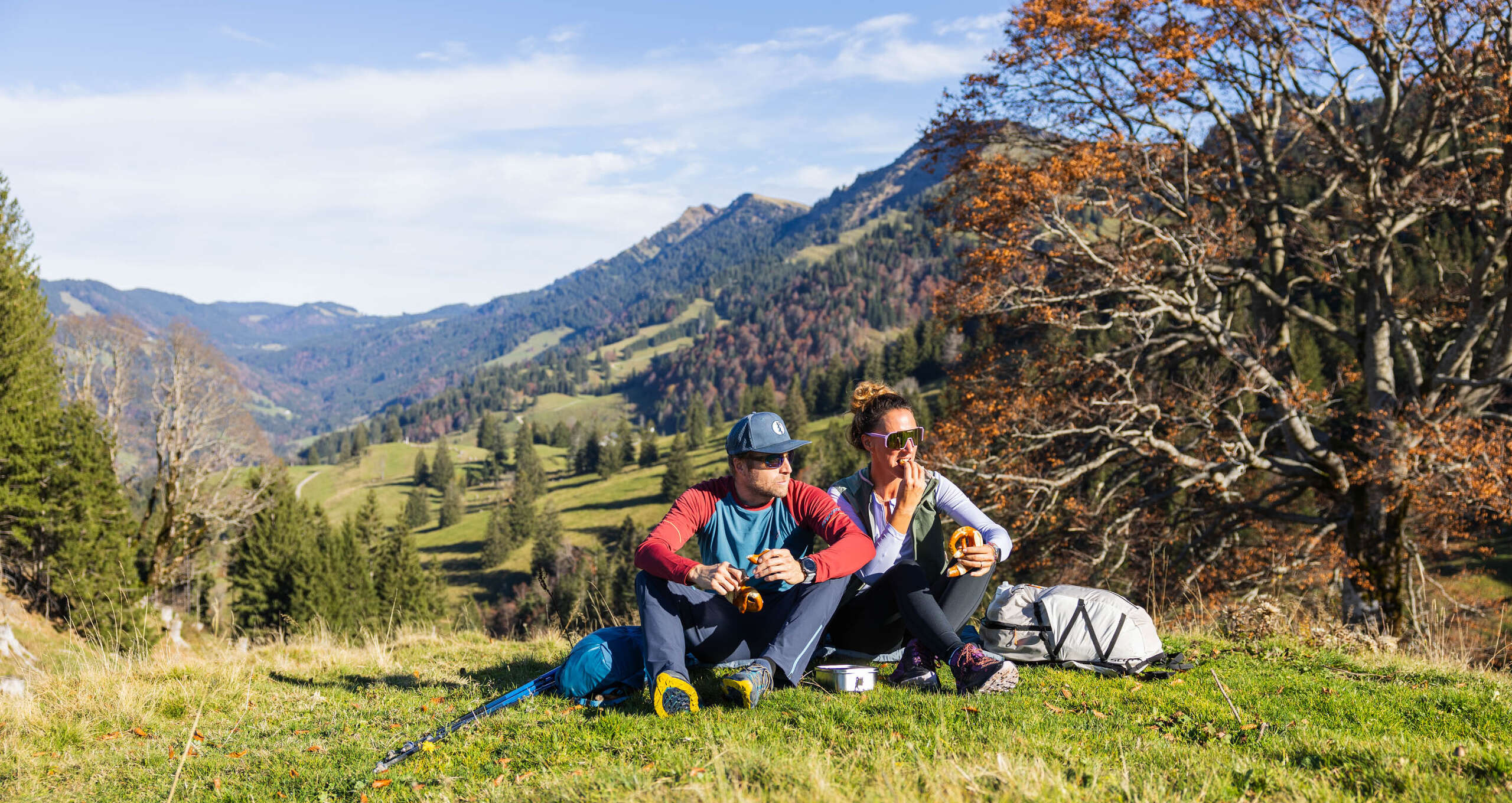 Wandern im Herbst bei Oberstaufen im Allgäu Ein Pärchen sitzt auf der Kuppe und genießt die Sonne. Im Hintergrund sind Berge und herbstliche Wälder zu sehen.