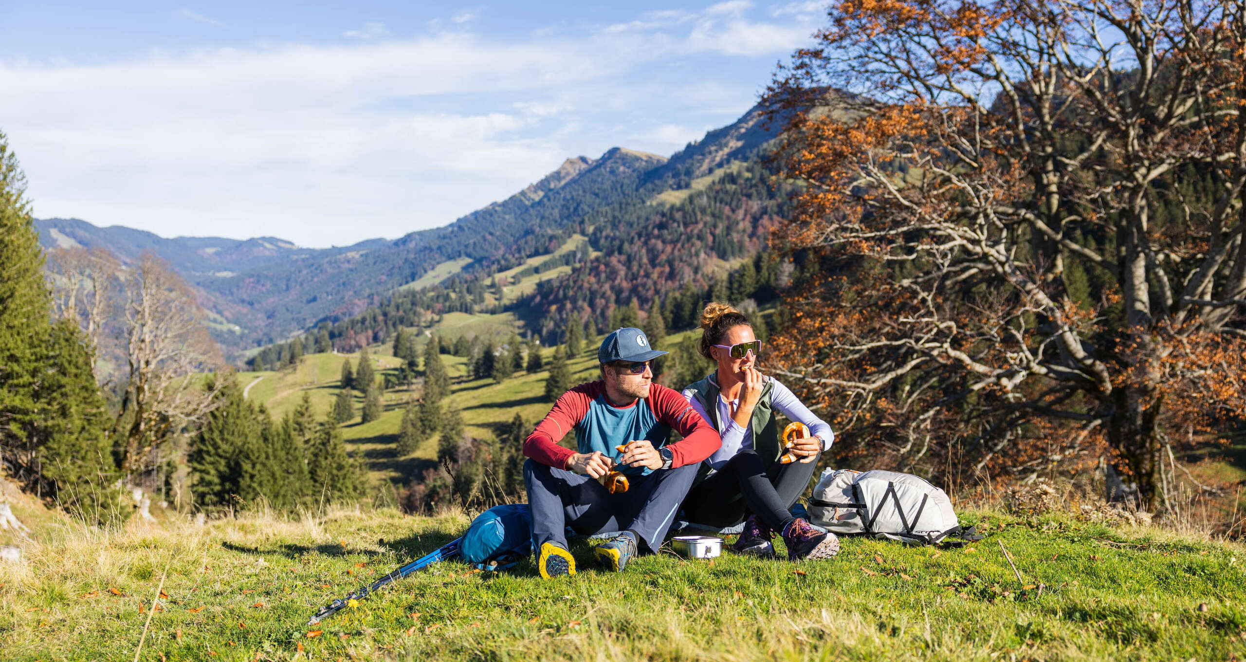 Wandern im Herbst bei Oberstaufen im Allgäu Ein Pärchen sitzt auf der Kuppe und genießt die Sonne. Im Hintergrund sind Berge und herbstliche Wälder zu sehen.