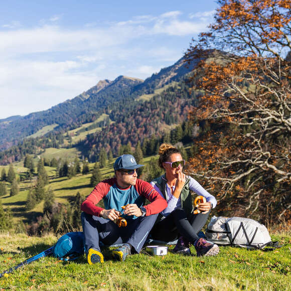 Ein Pärchen sitzt auf der Kuppe und genießt die Sonne. Im Hintergrund sind Berge und herbstliche Wälder zu sehen.